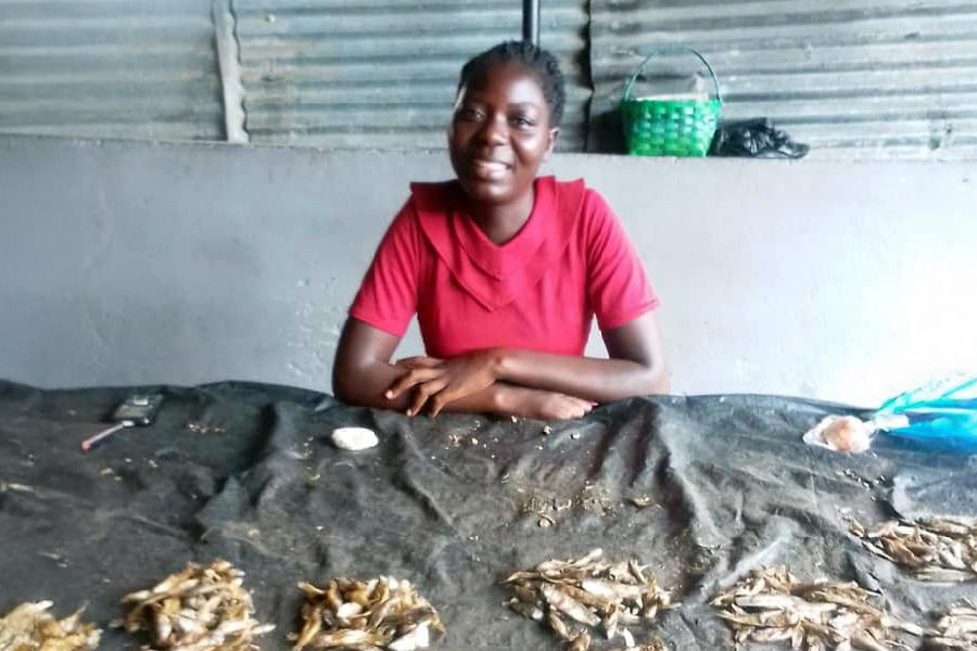 A young Zambian woman wearing a red t-shirt stands beside her fish counter. 