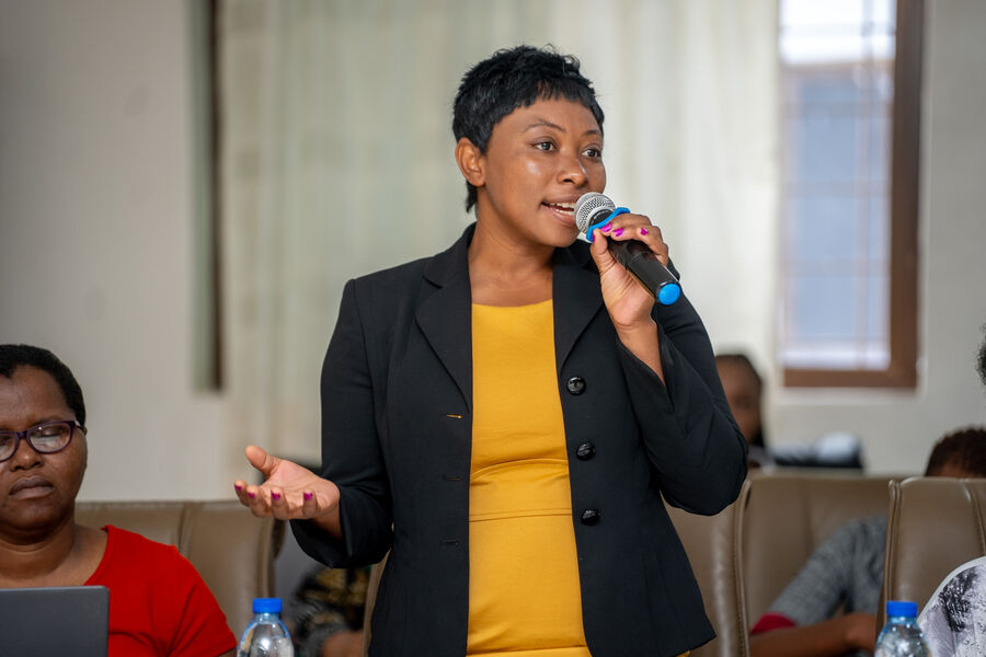A Zimbabwean woman with short hair and wearing a yellow dress and black jacket, speaks into a microphone.