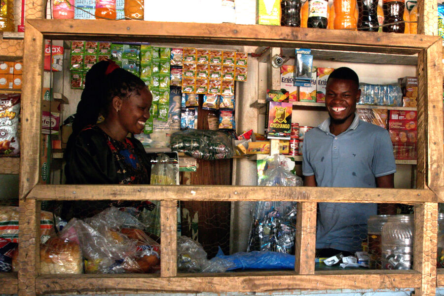 A young Malawian woman and man stand inside a grocery shop, smiling, surrounded by groceries.