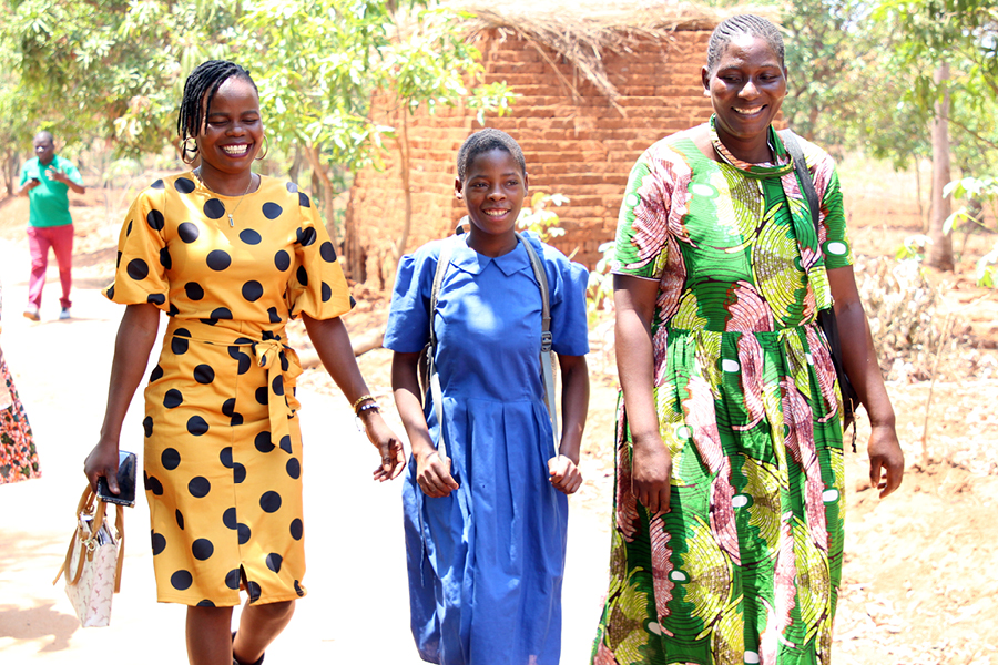 Two Malawian women wearing patterned dresses walk with a primary school student back to school.
