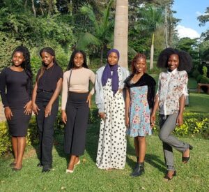 A group of six smiling women stand in a line for a photo outside. They are standing on grass, in front of trees.