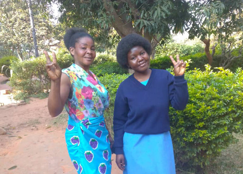 Jessica, a Zambian student in blue school uniform, with her mother who is wearing a colorful patterned dress. They are both smiling, throwing up peace signs with their hands, standing outside in front of green bushes and trees for a photo.