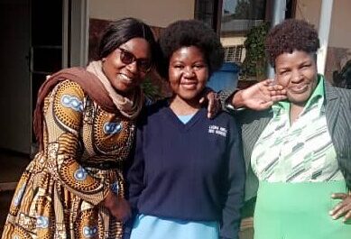 Jessica, a Zambian student in blue school uniform, standing between her Teacher Mentors Ireen (left) and Juliet. They are all smiling for a photograph stood outside the school.