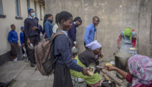 A Zimbabwean schoolgirl in uniform with a rucksack smiles as a member of a mother support group serves her a school meal.