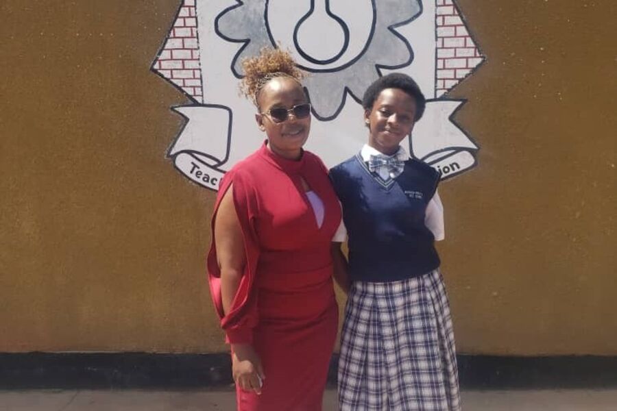 Hope, a smiling Zambian student in school uniform, stands beside her Teacher Mentor wearing a pink dress and sunglasses. They are in front of a yellow wall with a school logo.