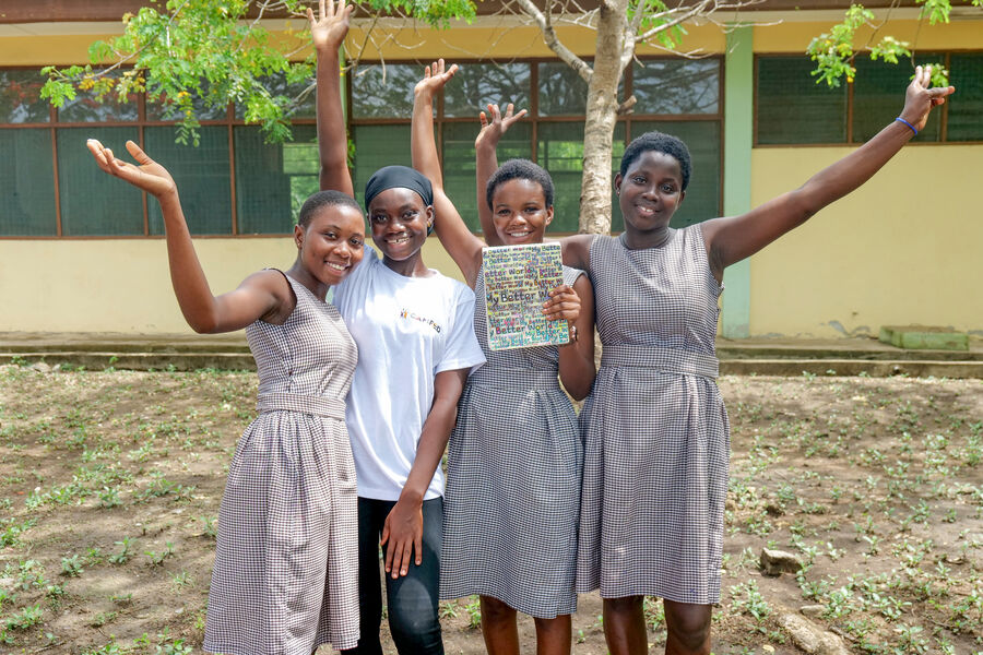 Innocent, a Ghanaian secondary school student in uniform, stands smiling with her Learner Guide Sandra, who is wearing a white CAMFED T-shirt and black headscarf, alongside two other girls in school uniform. All four raise their arms in celebration while standing outdoors, with trees and a school building visible in the background.
