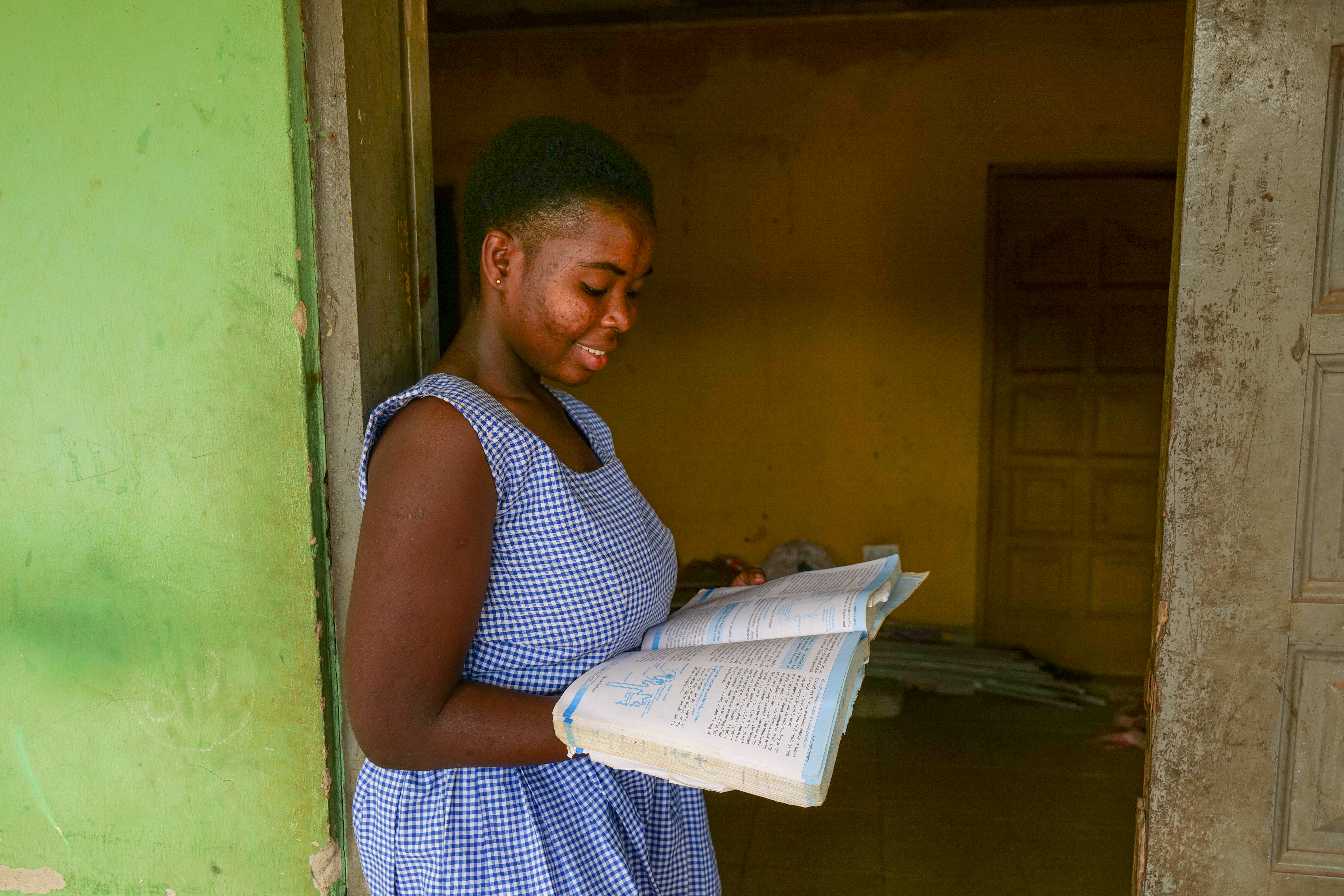 Ellen, a smiling Ghanaian student in a blue school dress, leans against a doorframe while reading an open textbook she holds in her hands.