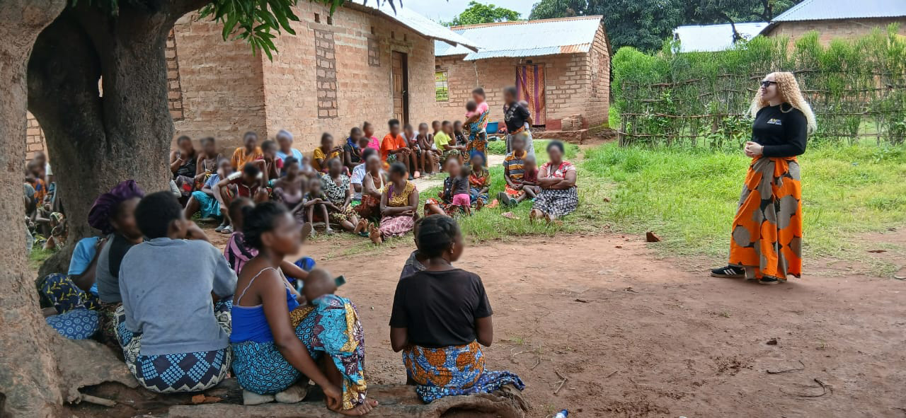 Gift (right), a Zambian woman in black top and patterned orange skirt, leads a community awareness session outside in Zambia.